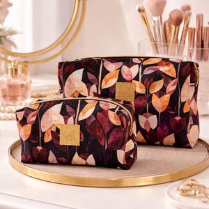 Può includere: Tre trousse per cosmetici con sfondo scuro e motivo a foglie nei toni del bordeaux, pesca e crema. Le trousse hanno cerniere dorate e un dettaglio quadrato dorato. Sono esposte su un vassoio dorato.