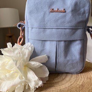 Könnte beinhalten: Eine hellblaue Canvas-Umhängetasche mit einem roségoldenen Reißverschluss und einem roségoldenen Metalletikett mit der Aufschrift "Handmade". Die Tasche liegt auf einer Holzoberfläche mit einer weißen Blume.