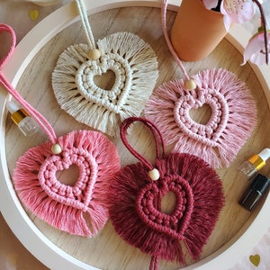 Könnte beinhalten: Vier handgefertigte Makramee-Herzornamente mit Fransen in Weiß, Rosa und Burgunderrot, die an einem weißen runden Holztablett hängen.