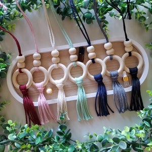 May include: A collection of wooden aromatherapy car air fresheners with colorful tassels. Each air freshener features a wooden ring, beads, and a tassel in various colors, including burgundy, pink, cream, mint, navy, gray, and black. The air fresheners are displayed on a white tray.