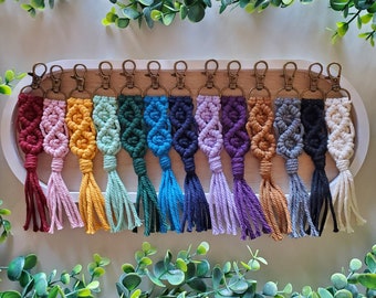 May include: A collection of macrame keychains in various colors, including red, pink, yellow, green, blue, purple, brown, gray, black, and cream. Each keychain features a woven design and a tassel, attached to a bronze-colored clasp.