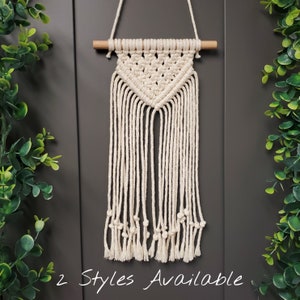 Peut inclure: Une suspension murale en macramé blanche avec un motif triangulaire et des franges. La suspension est fixée à une baguette en bois.