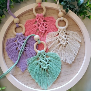 Puede incluir: Cuatro llaveros de macramé en forma de hoja con anillos de madera, cada uno en un color diferente: rosa, morado, verde y crema.