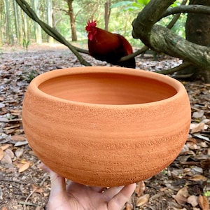 May include: A round, terracotta-colored ceramic bowl with a textured, matte exterior surface featuring horizontal ridges. The bowl has a wide, open top and a smooth, lighter-toned interior rim, showcasing a natural, earthy clay finish throughout.