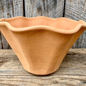 May include: A terracotta-colored, handmade flower pot with a unique, wavy rim. The pot has a geometric, faceted design and a natural, earthy texture. The pot is suitable for indoor or outdoor use.