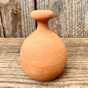 Op de afbeelding: Een kleine, ongeglazuurde terracotta vaas met een afgeronde body, een smalle hals en een platte bovenkant. De vaas heeft een getextureerd oppervlak en een warme, aardse oranje kleur. De vaas is waarschijnlijk handgemaakt en ongeveer 10 cm hoog.