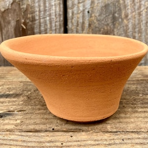 May include: A small, unglazed terracotta pot with a flared, conical shape. The pot has a wide opening and a textured surface. The color is a warm, earthy orange-brown. The pot is sitting on a weathered wooden surface.