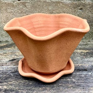 May include: A terracotta flower pot with a wavy, scalloped rim and a matching saucer. The clay vessel features a textured, matte surface in a natural reddish-brown hue, with a wide opening that tapers down to a smaller, circular base.