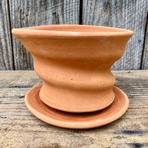 Puede incluir: Una maceta decorativa de color terracota con un diseño retorcido único. La maceta se asienta sobre un platillo a juego, ambos hechos de arcilla sin esmaltar. El diseño presenta una parte superior ancha y acampanada y un cuerpo esculpido y curvado.
