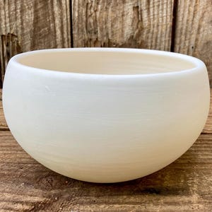 Könnte beinhalten: Eine runde, cremefarbene Keramikschale mit glatter, matter Oberfläche. Die Schale hat eine weite Öffnung und einen abgerundeten Boden. Der Hintergrund ist eine Holzoberfläche.