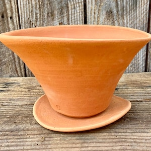 Puede incluir: Una maceta acampanada de color terracota con un platillo a juego. La maceta tiene una parte superior ancha y abierta y una forma cónica suavemente inclinada. El platillo es un plato redondo simple. Los artículos están hechos de arcilla sin esmaltar.