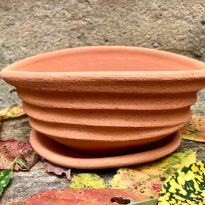 May include: A terracotta plant pot with a wide, open top and a matching saucer. The exterior of the bowl features a series of horizontal, raised ridges that wrap around the circumference, creating a textured, ribbed appearance in a natural clay color.