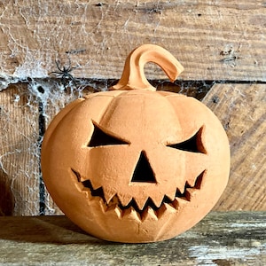 May include: A carved terracotta pumpkin jack-o'-lantern with a matte orange finish. The pumpkin features a curved stem at the top and cut-out facial features including two triangular eyes and a wide, jagged mouth with sharp, pointed teeth.