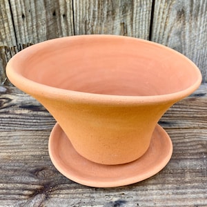 Puede incluir: Una maceta acampanada de color terracota con un platillo a juego. La maceta tiene una parte superior ancha y abierta y una forma suavemente curvada. El platillo es redondo y se encuentra debajo de la maceta, proporcionando una base estable. La maceta y el platillo son de arcilla sin esmaltar.