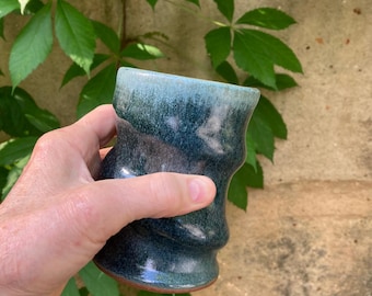May include: A handmade ceramic mug with a unique, wavy design. The mug features a blend of teal and dark blue hues, with a lighter blue rim. The base has a reddish-brown color. The mug is held in a hand, with green leaves in the background.