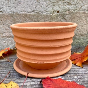 Puede incluir: Una maceta de color terracota con diseño acanalado y platillo a juego. La maceta tiene una parte superior ancha y abierta y una serie de crestas horizontales. La maceta está sobre una superficie de madera con hojas de otoño coloridas.