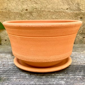 Puede incluir: Una maceta redonda de color terracota con un platillo a juego. La maceta tiene una forma ancha y ligeramente acampanada con dos ranuras horizontales cerca de la parte superior. El platillo es un plato simple y poco profundo. La maceta mide unos 20 cm de diámetro.