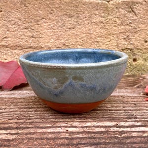 Puede incluir: Un cuenco de cerámica hecho a mano con interior azul y exterior texturizado gris claro. La base es de color marrón anaranjado cálido. El cuenco está sobre una superficie de madera, con una hoja roja visible al lado.