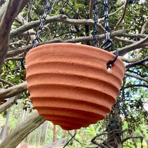May include: A terracotta hanging planter with a ribbed, horizontal texture. The bowl-shaped pot features three evenly spaced holes along the upper rim, which are connected to a black metal chain system for suspension from a sturdy support.