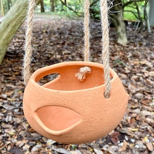 Op de afbeelding: Een ronde, terracotta-kleurige hangende plantenbak met een getextureerd oppervlak. De komvormige houder heeft twee grote, ovale openingen aan de zijkanten en wordt opgehangen aan drie dikke, natuurlijk gekleurde, gedraaide jutekoorden.