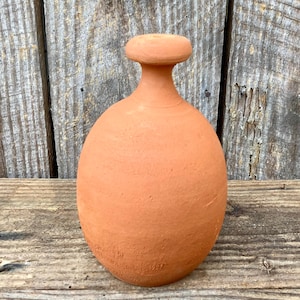 Op de afbeelding: Een terracotta-kleurige, afgeronde kleifles met een korte hals en een platte, cirkelvormige bovenkant. De fles heeft een glad, ongeglazuurd oppervlak en staat op een houten oppervlak. De achtergrond is een houten muur.