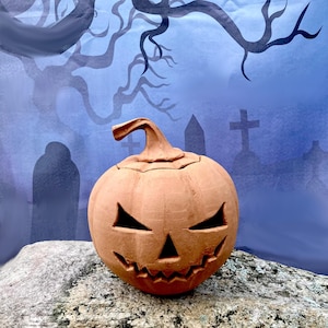 May include: A terracotta-coloured ceramic pumpkin jack-o'-lantern featuring a carved, jagged, triangular-eyed face with a wide, toothy grin and a curved stem on top. The pumpkin has a matte, earthy finish and is shaped with vertical ridges.