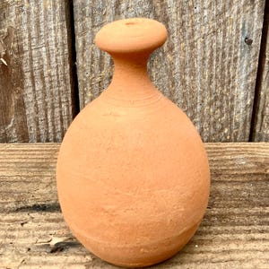 Op de afbeelding: Een terracotta-kleurige, bolvormige vaas met een smalle hals en een platte, ronde bovenkant. Het handgemaakte aardewerk heeft een textuur en wordt tentoongesteld tegen een verweerde houten achtergrond. De vaas wordt waarschijnlijk gebruikt voor decoratieve doeleinden.