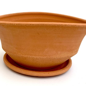 Puede incluir: Una maceta de terracota de forma ovalada con un platillo a juego. La maceta tiene un borde ensanchado y un cuerpo curvado. El platillo es una base redonda simple. El diseño es sencillo y rústico, adecuado para varias plantas. Medidas en centímetros.