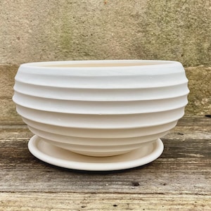 May include: A matte white ceramic planter bowl featuring a series of horizontal, raised ridges encircling the exterior. The bowl has a wide, open top and sits securely on a matching circular ceramic saucer base, creating a cohesive unit.