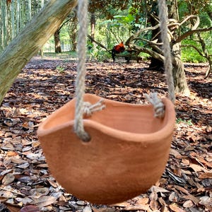 May include: A terracotta-colored clay hanging planter with a rounded, bowl-like shape and an uneven, wavy rim. The planter is suspended by two thick, light-colored twisted jute ropes that are knotted through holes on opposite sides of the rim.