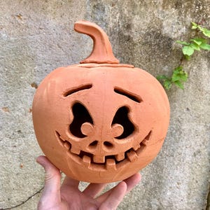 May include: A terracotta-coloured ceramic pumpkin sculpture featuring a carved jack-o'-lantern face with arched eyes, a small nose, and a wide, toothy grin. The pumpkin has a curved stem on top and a smooth, matte finish throughout the surface.