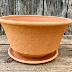 May include: A terracotta-colored, oval-shaped planter with a matching saucer. The planter has a wide, flared rim and a textured surface. The saucer is a smaller, circular base that fits beneath the planter. The background is a weathered wooden surface.