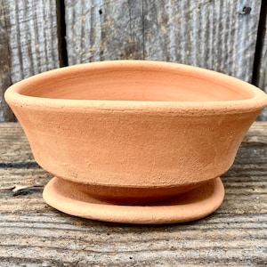 Puede incluir: Una pequeña maceta redonda de color terracota, con forma de cuenco ancho y poco profundo. La maceta tiene un borde ligeramente ensanchado y se asienta sobre una pequeña base circular. La textura es áspera y sin esmaltar.