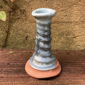May include: A handmade ceramic candle holder with a spiral design in shades of blue and gray. The candle holder sits on a terracotta-colored base. The top has a wide opening. The piece is displayed on a wooden surface.