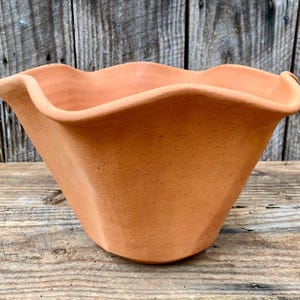 May include: A terracotta-colored, handmade planter with a unique, wavy rim. The planter has a square base that tapers upwards to a wider opening. The textured surface adds to its rustic charm. Perfect for indoor or outdoor use.