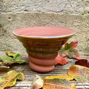 Op de afbeelding: Een handgemaakte keramische kom in terracotta kleur met een brede, licht uitlopende rand. De kom heeft een spiraalvormige basis en een geglazuurde, bruine en groene buitenkant. De kom is omgeven door kleurrijke herfstbladeren op een houten oppervlak.