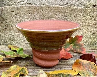 May include: A terracotta-coloured ceramic bowl with a wide, slightly flared rim. The bowl has a spiral-shaped base and a glazed, brown and green exterior. The bowl is surrounded by colourful autumn leaves on a wooden surface.
