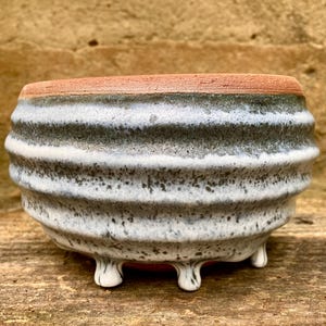 May include: A small, round ceramic bowl with a ribbed exterior texture. The bowl features a speckled white and grey glaze over a terracotta base, with a visible unglazed terracotta rim and three small, rounded feet at the base of the vessel.