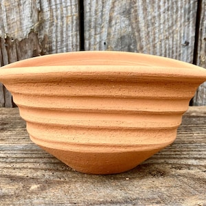 May include: A terracotta-colored, ribbed planter with a wide, flared rim. The planter has a rounded, conical shape and is made of unglazed clay. The horizontal ridges create a textured surface. The background is a weathered wooden surface.
