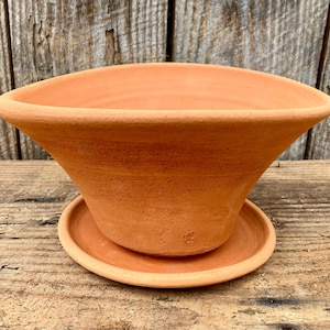 Puede incluir: Una maceta acampanada de color terracota con un platillo a juego. La maceta tiene una parte superior ancha y abierta y un exterior texturizado suavemente inclinado. El platillo se encuentra debajo de la maceta, proporcionando una base estable. El diseño rústico es adecuado para uso en interiores o exteriores.