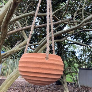 May include: A terracotta-colored hanging planter with a rounded, ribbed exterior texture. The bowl-shaped vessel features three evenly spaced holes along the upper rim, through which a thick, natural-colored jute rope is threaded for suspension.