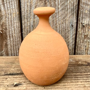 Op de afbeelding: Een terracotta-kleurige, afgeronde vaas met een smalle hals en een platte, schijfvormige bovenkant. De vaas heeft een glad, ongeglazuurd oppervlak en staat op een houten oppervlak. De vaas is waarschijnlijk handgemaakt en heeft een rustieke uitstraling.