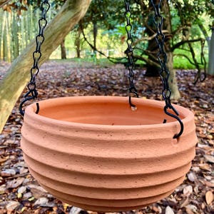 May include: A round terracotta hanging planter with a ribbed, horizontal texture on the exterior. The bowl-shaped pot features three evenly spaced holes along the upper rim, which are attached to a black metal chain for suspension purposes.