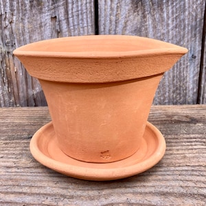 May include: A terracotta-colored flower pot with a matching saucer. The pot has a flared shape with a wide rim and a slightly curved top. The saucer is round and fits snugly beneath the pot. The pot and saucer are made of unglazed clay.