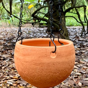 May include: A round, terracotta-colored hanging planter with a textured, porous surface. The bowl-shaped vessel features three evenly spaced circular holes along the upper rim, which are connected to a black metal chain for suspension purposes.