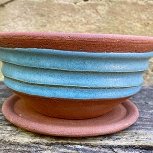 May include: A terracotta planter bowl with a matching saucer, featuring a wide, flared rim and three horizontal ridges around the exterior. The upper portion of the bowl is glazed in a matte light blue, while the base and saucer remain natural.