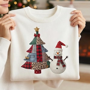 May include: White sweater featuring a Christmas tree and snowman design. The tree is made of patchwork fabrics, including leopard print and plaid. The snowman wears a red and white plaid scarf and hat. A gold star tops the tree.