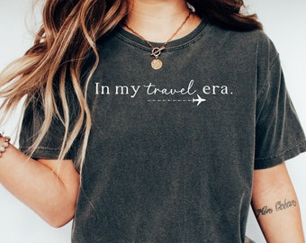 May include: A dark gray t-shirt with the text "In my travel era." and a small airplane graphic. The shirt is worn by a person with long blonde hair and a gold necklace.