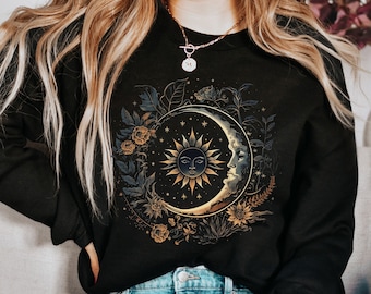 May include: Black sweatshirt with a gold sun and moon design surrounded by floral details. The sun and moon have faces and are set against a dark background.