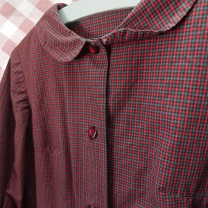 May include: A long-sleeved shirt featuring a small red and black checkered pattern. The garment includes a rounded Peter Pan collar and a front button placket with dark red buttons. The fabric has a fine texture and is displayed on a hanger.